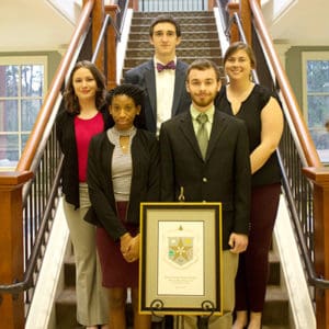 Five inductees into Kappa Mu Epsilon stand on stairs behind a plaque of the KME badge.