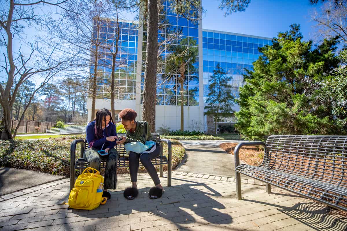 Two FMU students sit on a bench on a sunny day near the Leatherman Science Facility on FMU's campus.